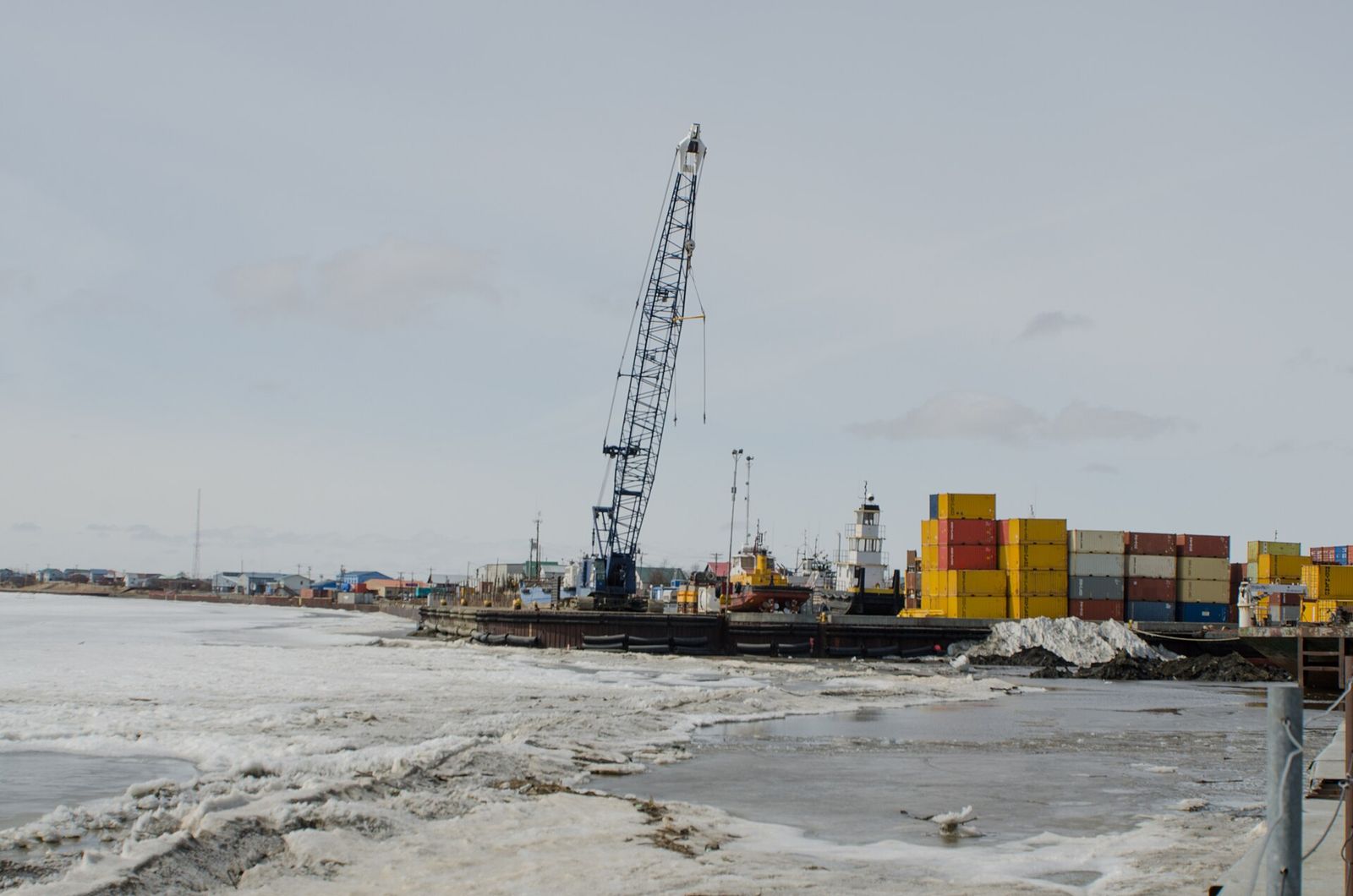 A crane and stacked shipping containers at the Bethel barge harbor, with ice still along the Kuskokwim River shore