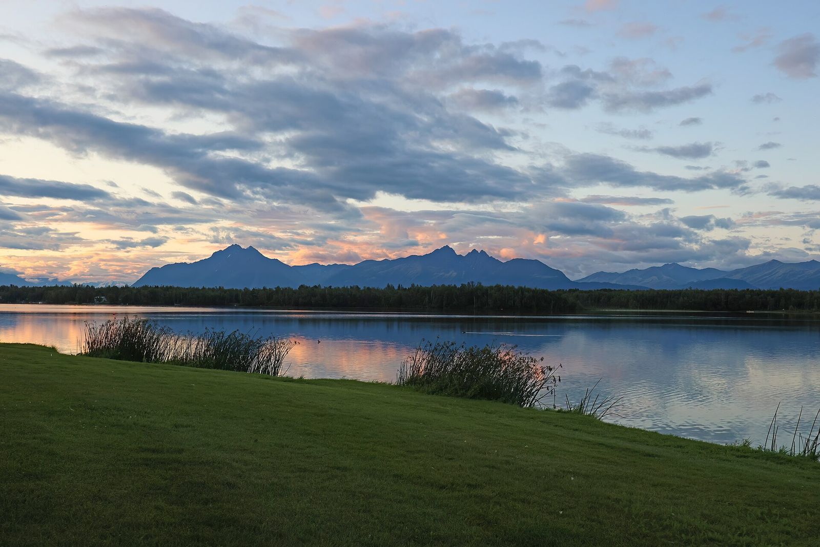 Morning light on Lake Lucille in Wasilla, with a grassy shoreline and mountains in the distance