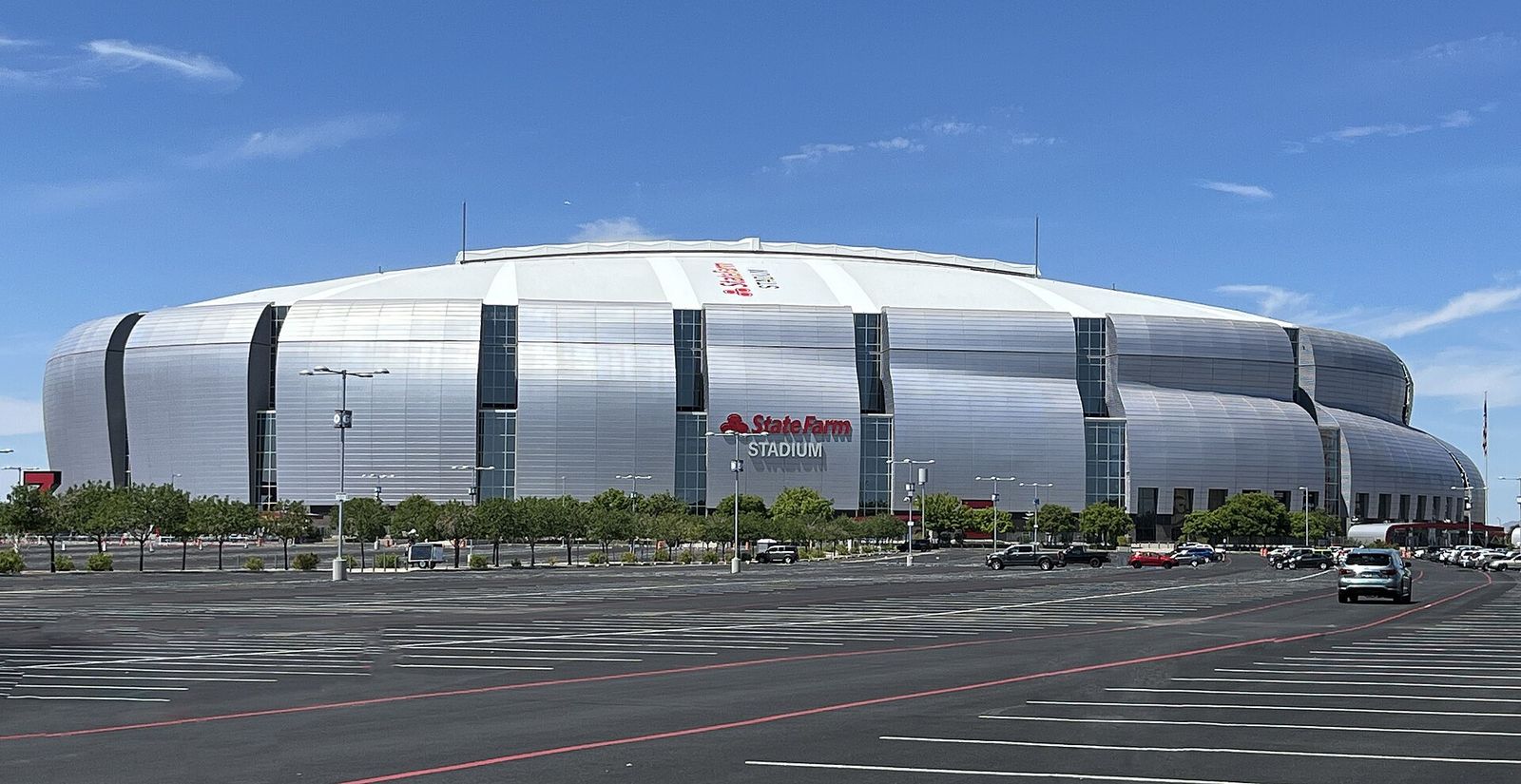 The silver curved exterior of State Farm Stadium in Glendale, seen across its parking lot under a blue sky