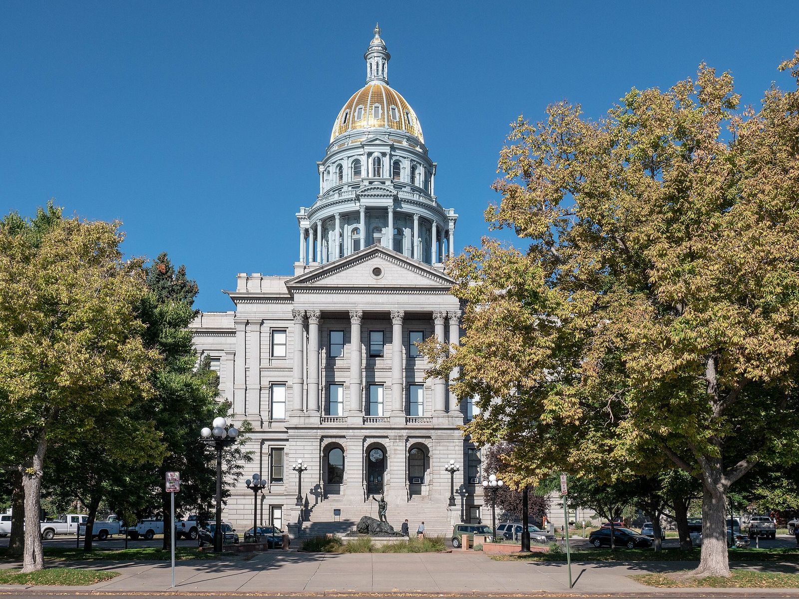 The Colorado State Capitol in Denver, with its gold dome and gray granite front, framed by autumn trees under a blue sky