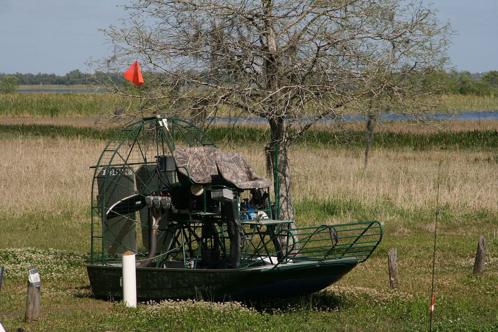 A Boggy Creek airboat with its tall caged fan parked on grass beside a bare tree at the edge of the marsh