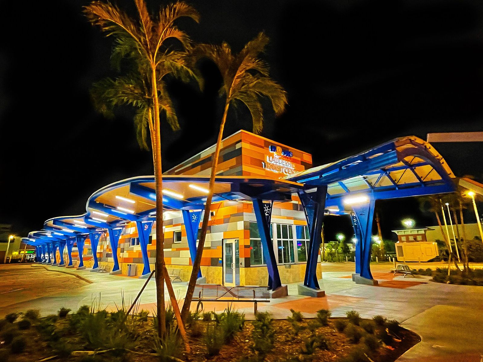 The Lauderhill Transit Center at night, its curved blue canopy and colorful building lit up beside palm trees
