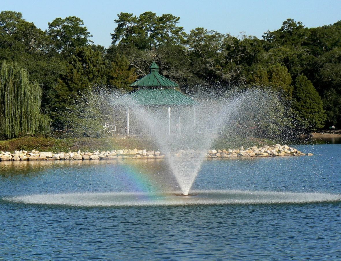 A fountain spraying in Lake Ella, with a green-roofed gazebo and trees behind it, in Tallahassee