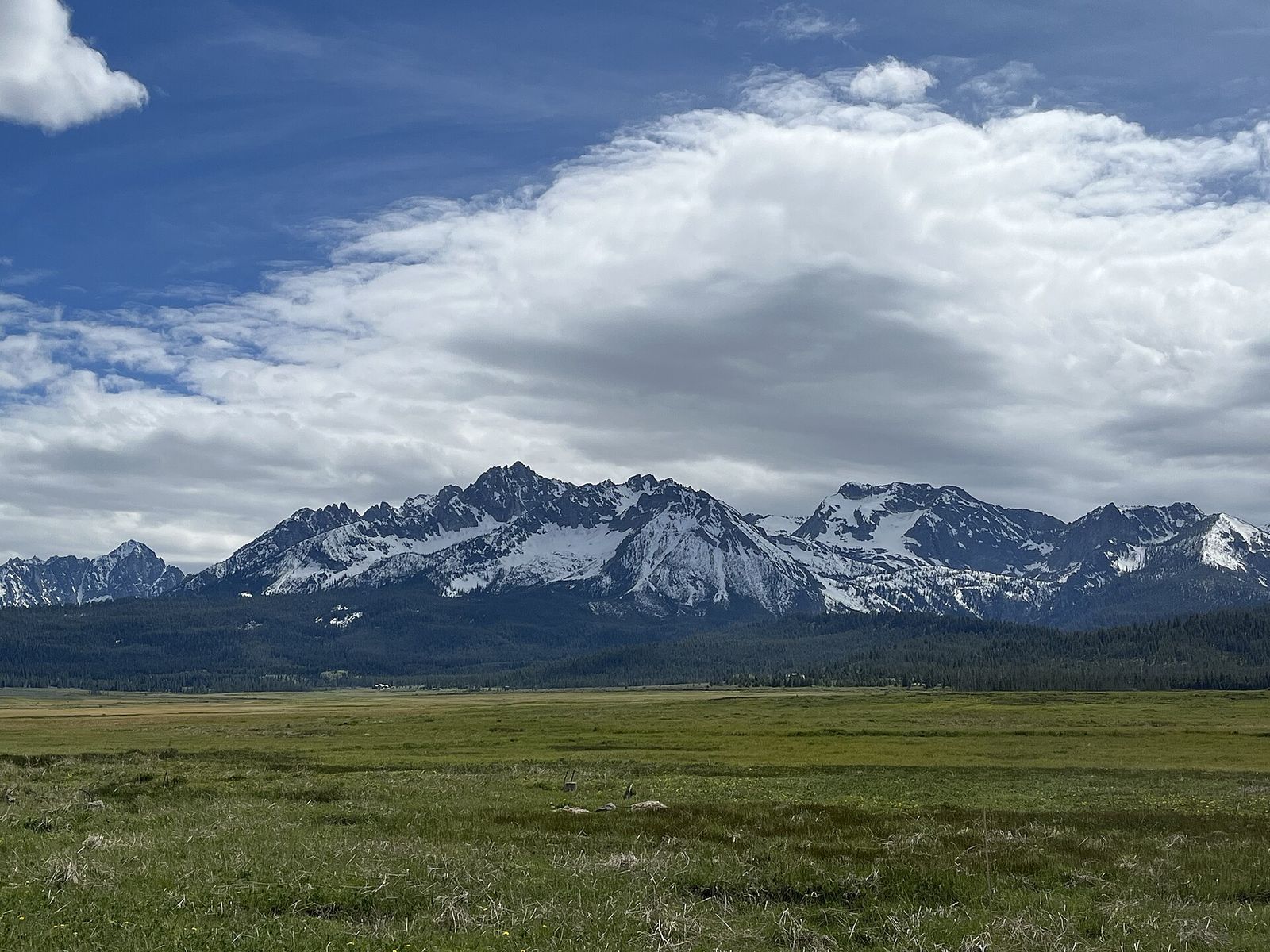 The jagged, snow-streaked Sawtooth Range rising beyond a wide green meadow under billowing clouds