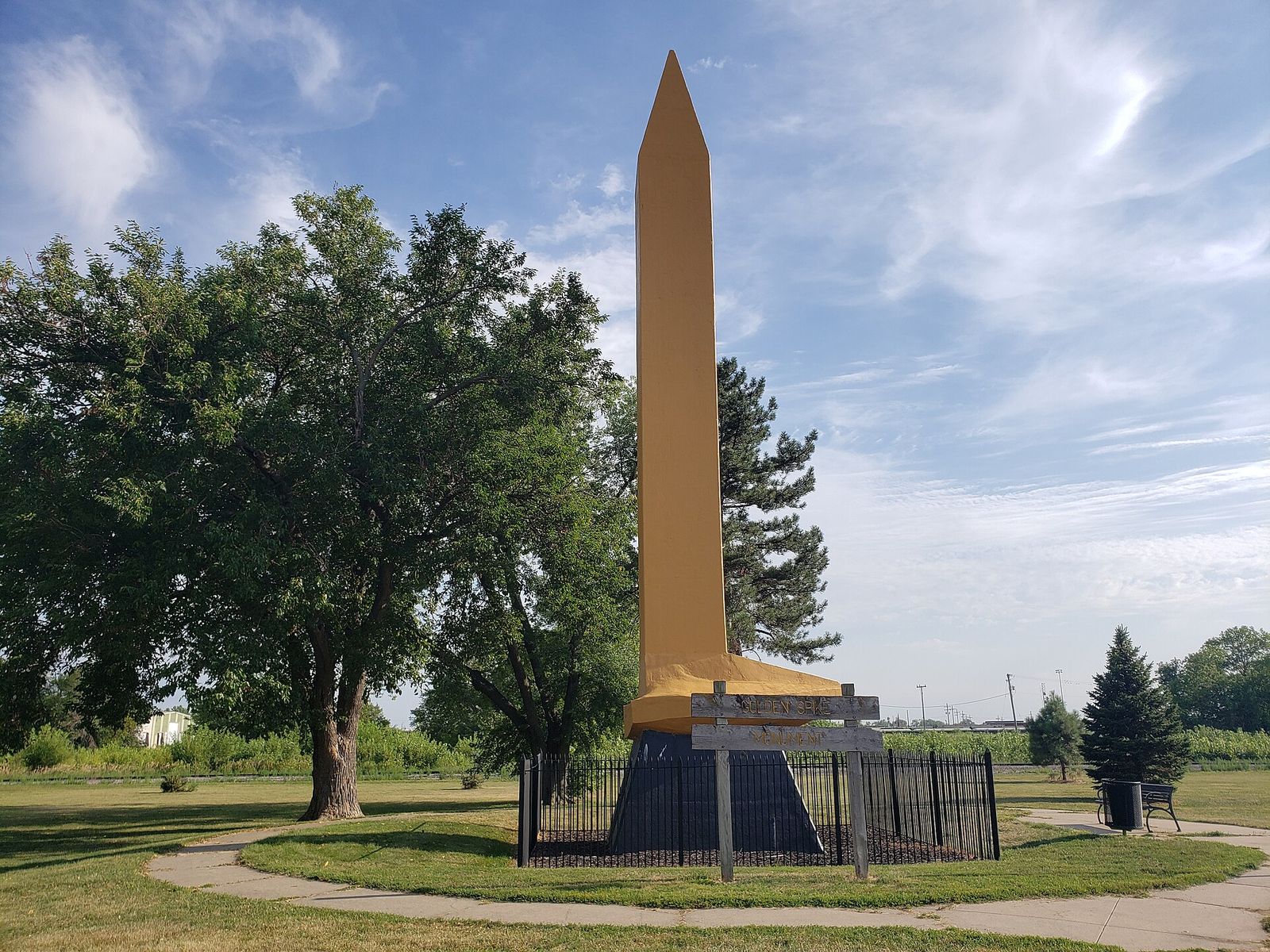 The tall gold-painted Golden Spike Monument in Council Bluffs, standing on a lawn beside leafy trees