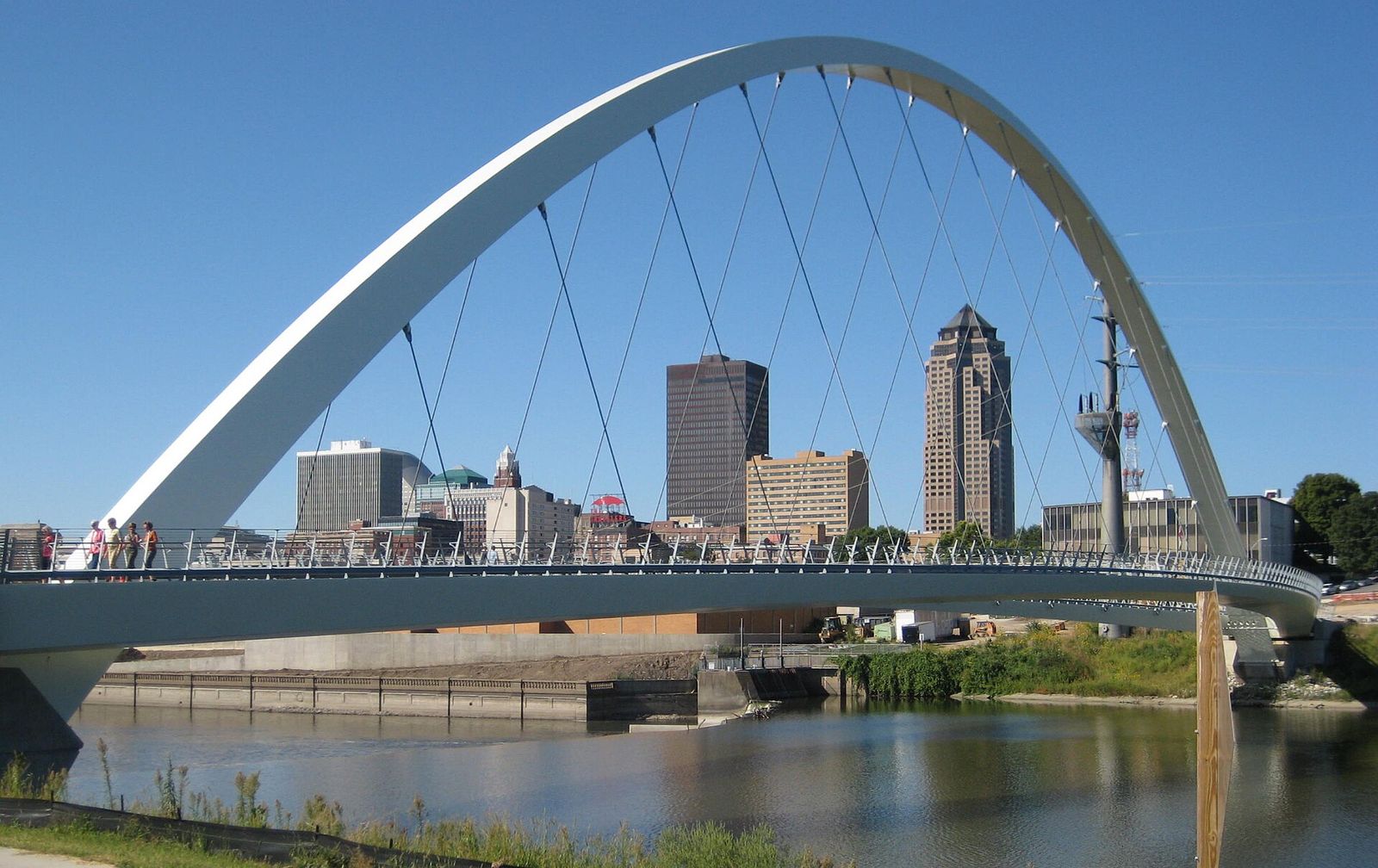 A white arched pedestrian bridge over the Des Moines River with the downtown Des Moines skyline behind it