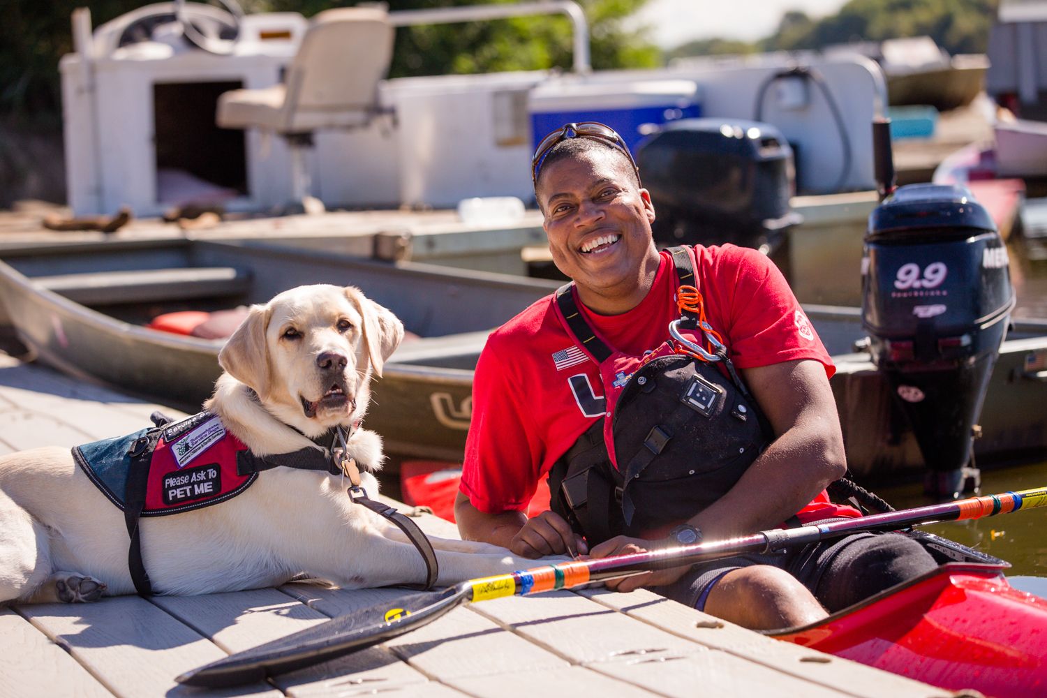 A yellow Labrador service dog in a vest lying on a dock beside its handler before a kayak training session