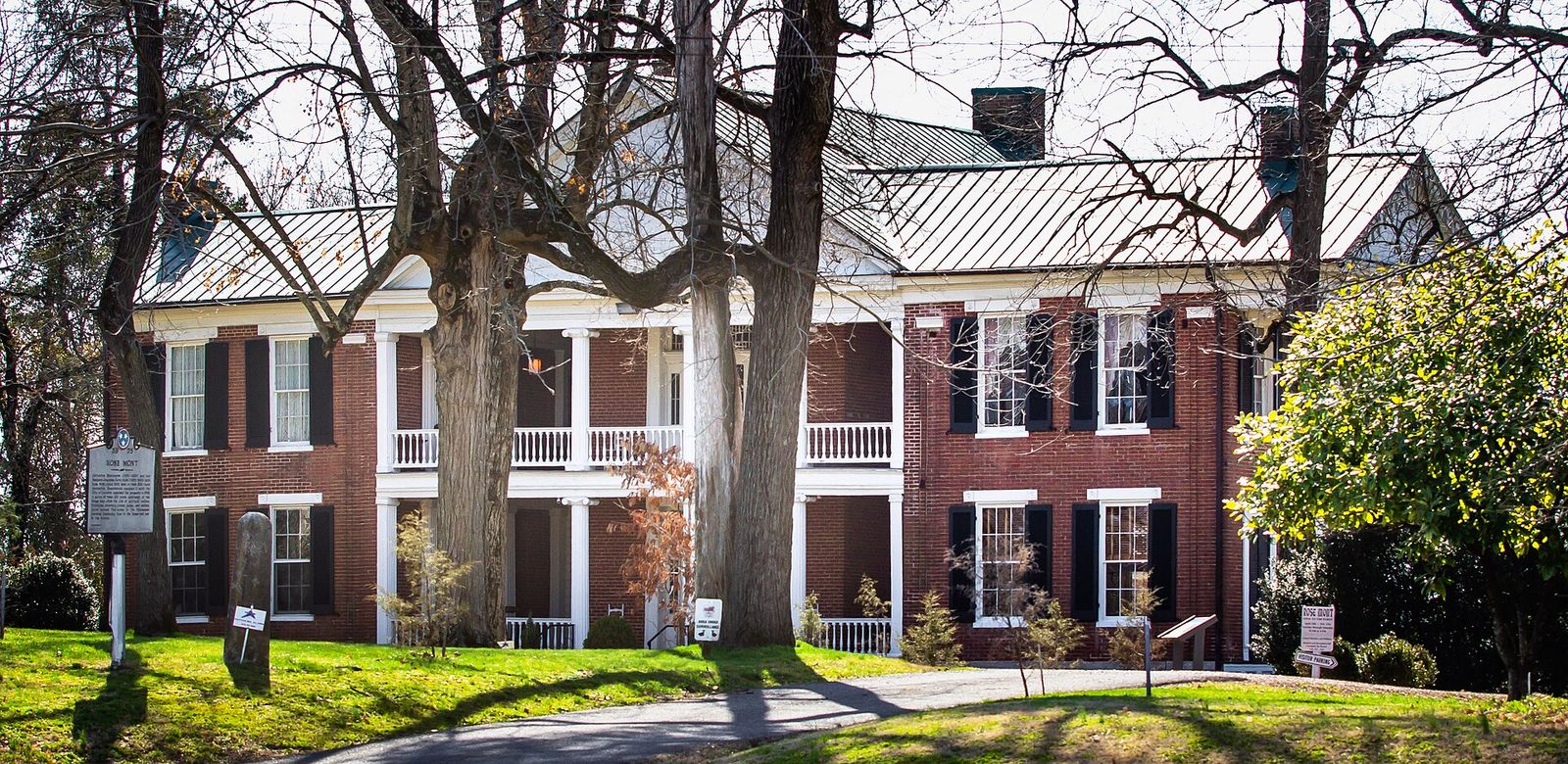 The brick Rosemont house with white porches behind large trees in Gallatin