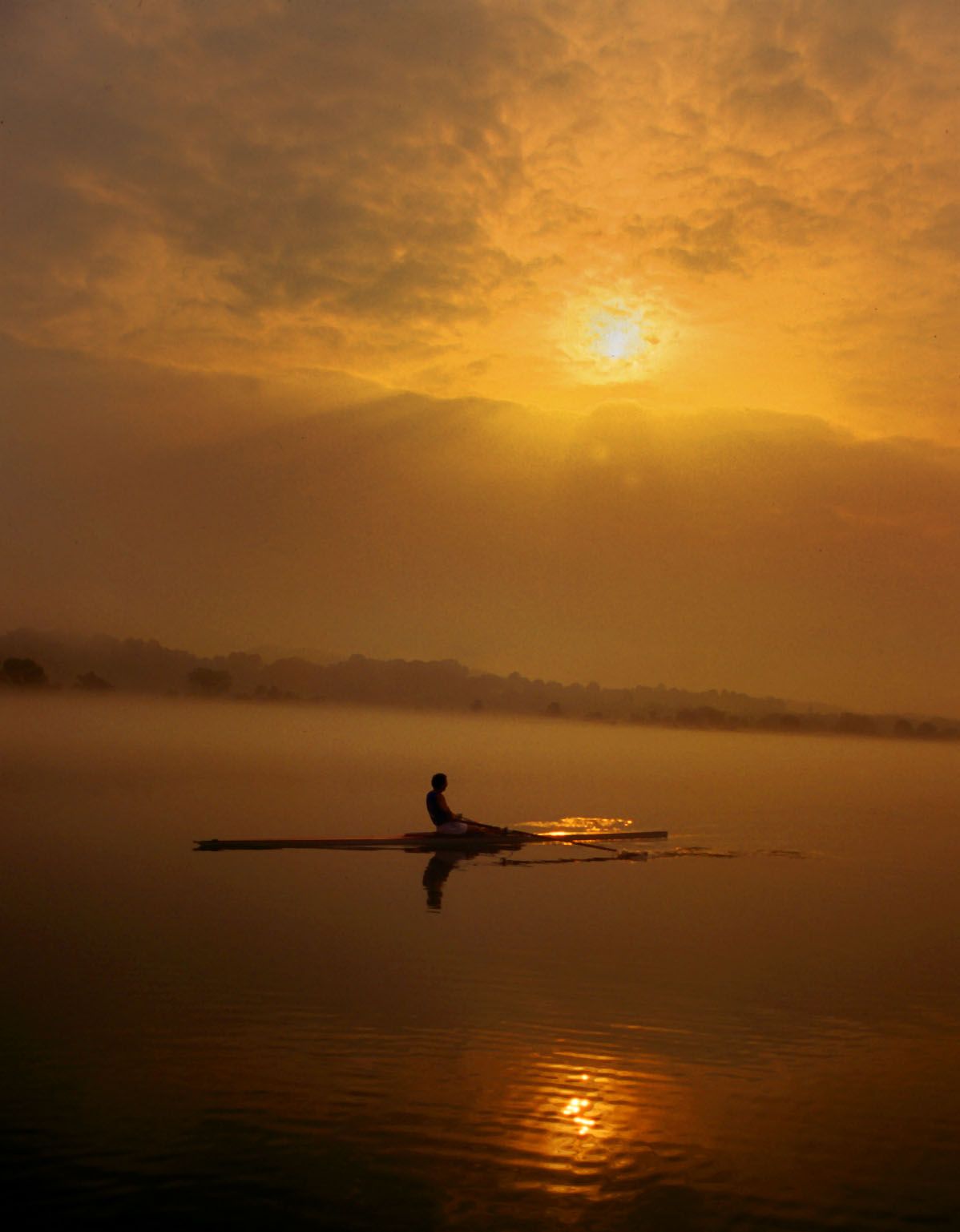 A single sculler on Melton Hill Lake under a hazy golden sunrise