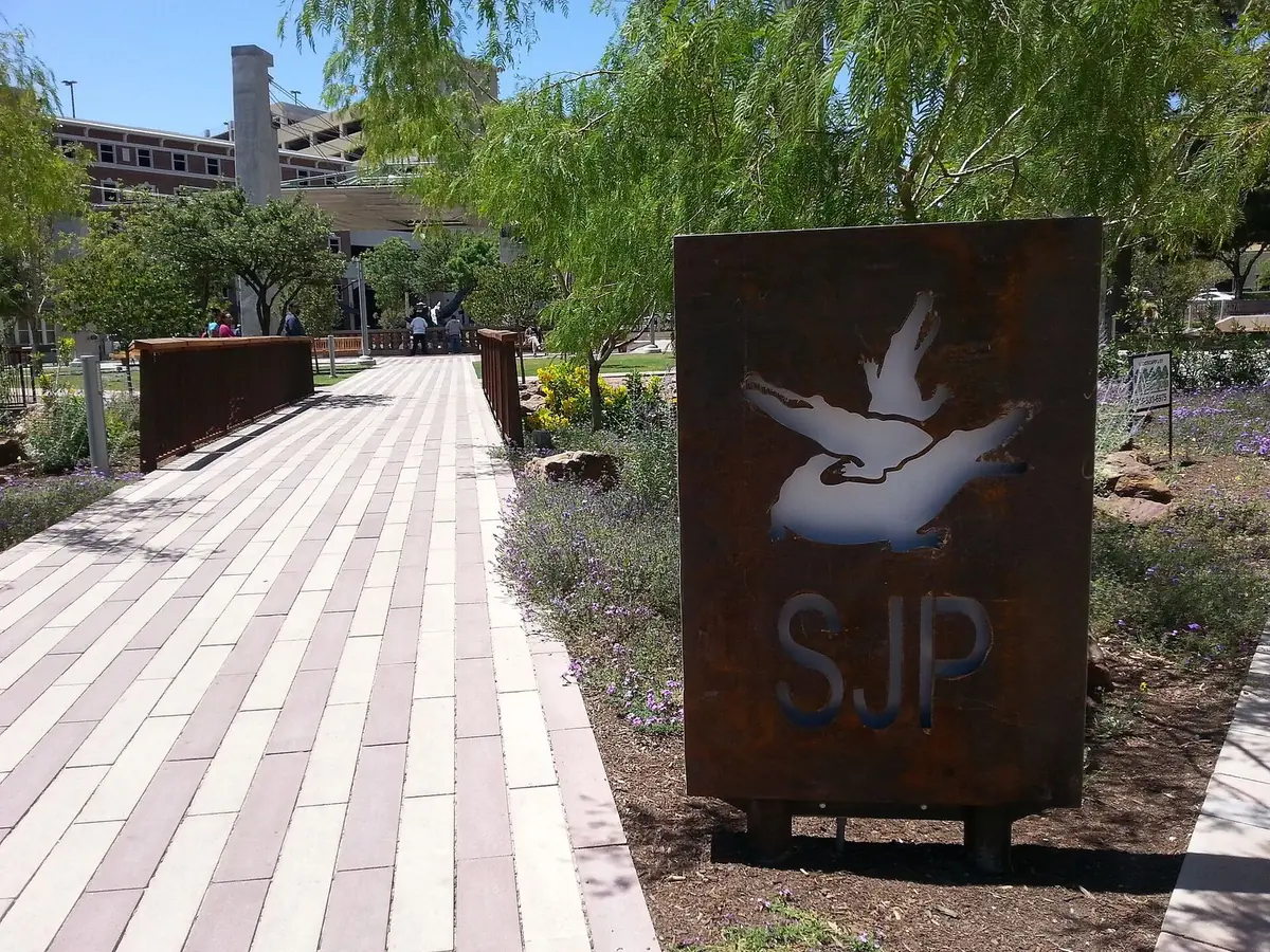 A paved walk leading into San Jacinto Plaza in downtown El Paso, beside the plaza's sign and shade trees
