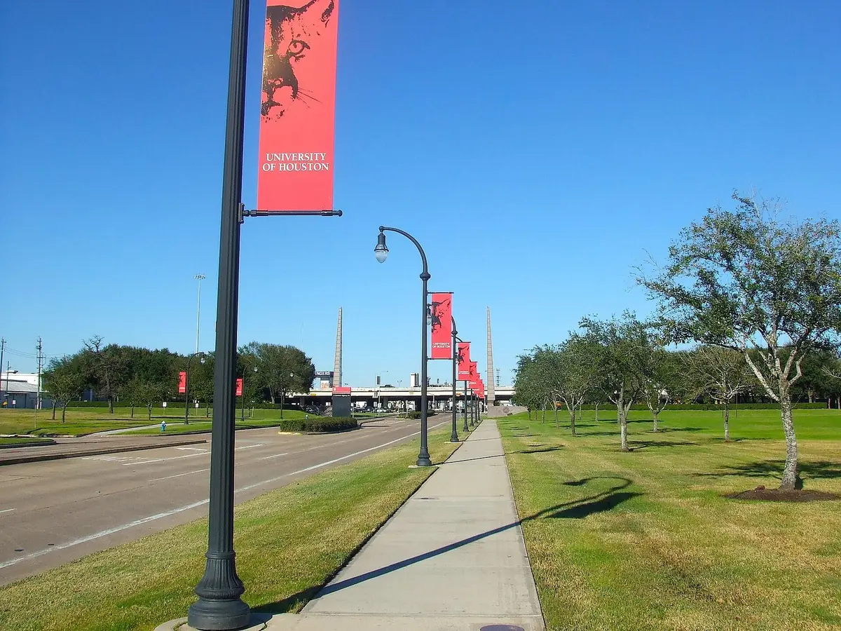 Red University of Houston banners on lamp posts along a sidewalk at a campus entrance on Cullen Boulevard