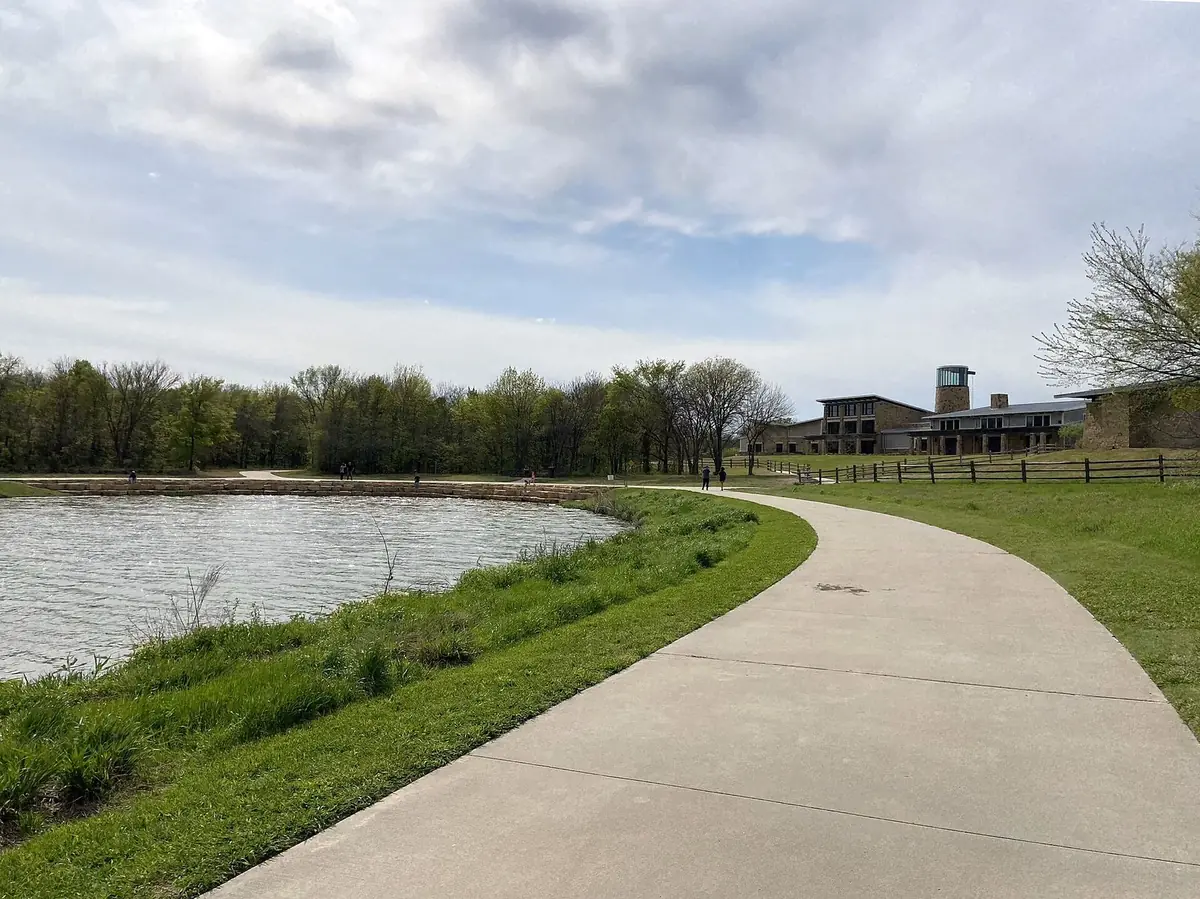 A paved path beside a pond leading to the Nature and Retreat Center at Oak Point Park in Plano