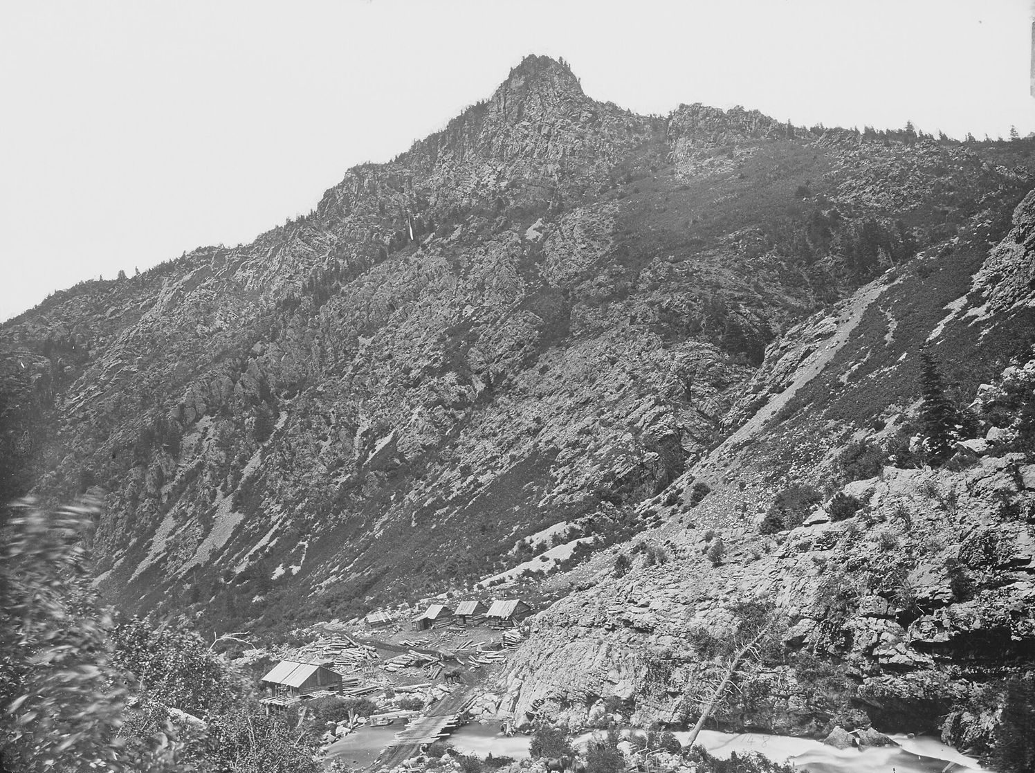 An early black and white photograph of American Fork Canyon with cabins below a rocky peak, by Timothy H. O'Sullivan