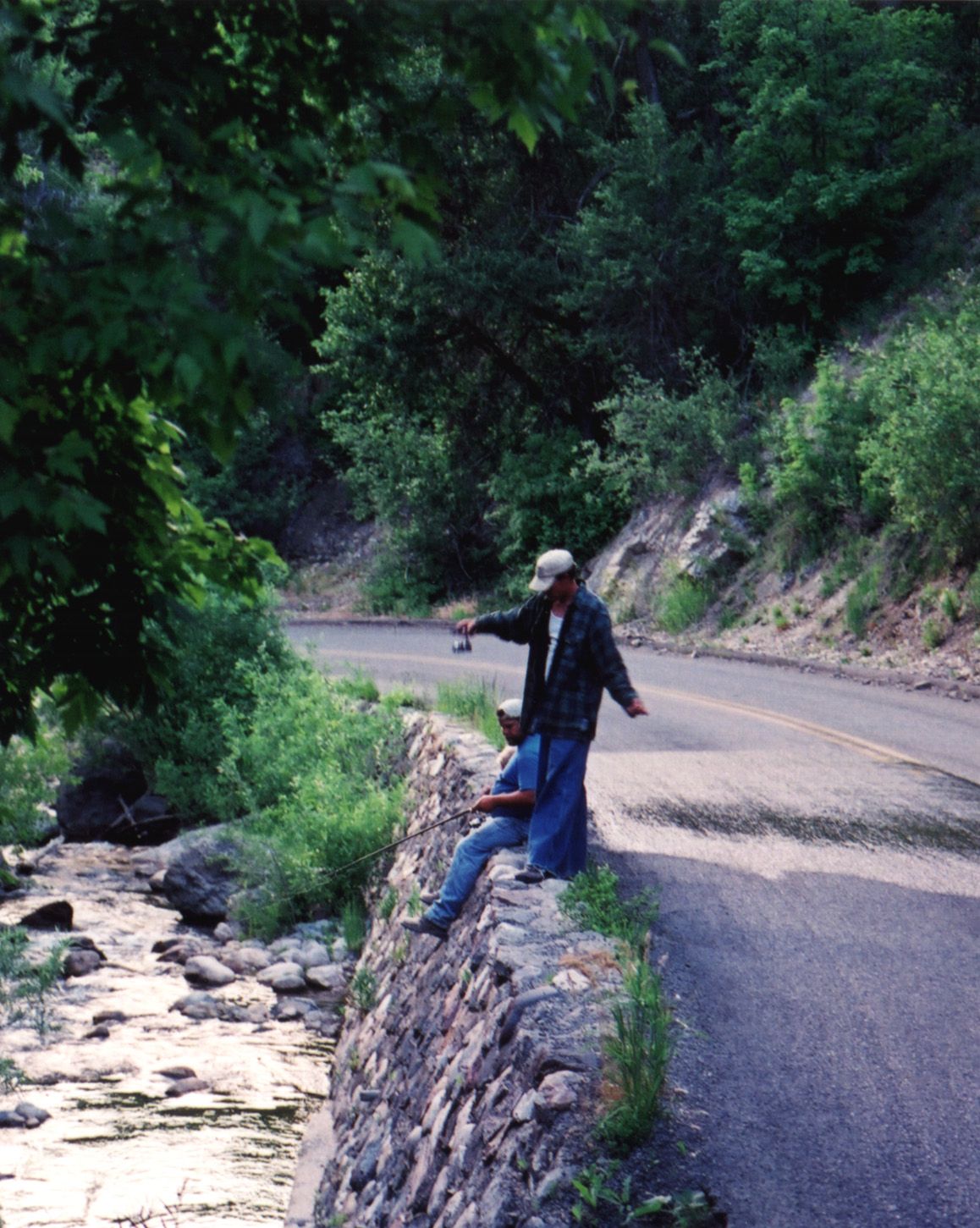 Two men fishing from a low wall of rounded stones beside Payson Creek, under trees in Payson Canyon