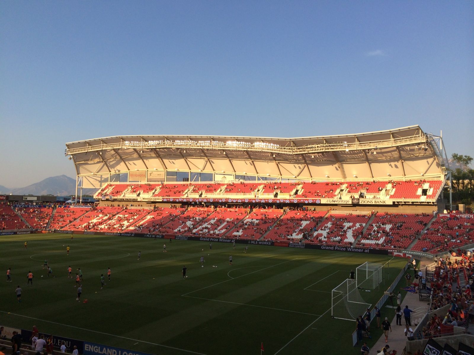 Players warming up on the pitch inside the Sandy soccer stadium, with red seats and a curved roof