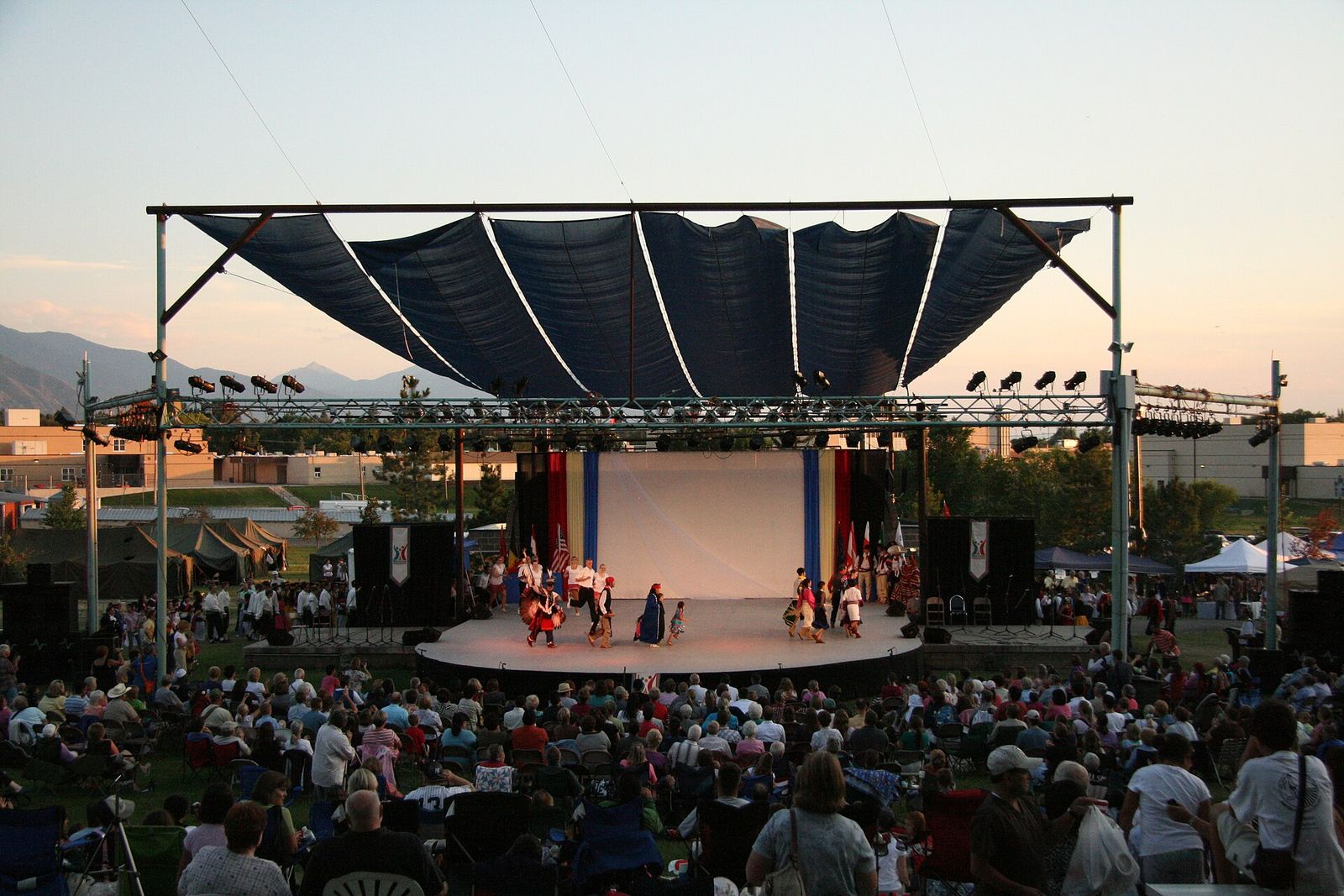 Performers on the covered outdoor stage at Springville World Folkfest, watched by a seated evening crowd