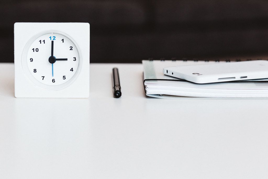 White analog clock beside a pen and notebook on a clean white desk