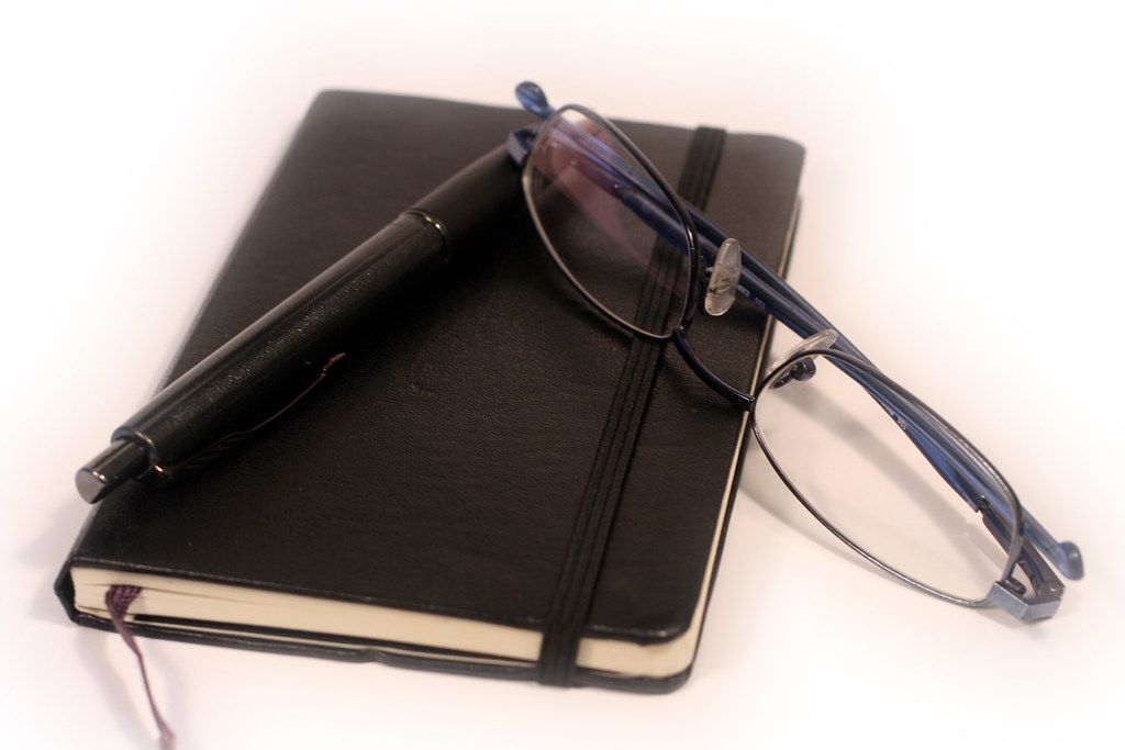 Black notebook with a pen and a pair of glasses resting on top